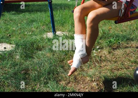 Kinderbeine sind mit Pflaster verbunden - barfuß auf einer Schaukel. Traumatische Ausrüstung, Kinderverletzung, Bluterguss, Verstauchung, Fraktur Stockfoto