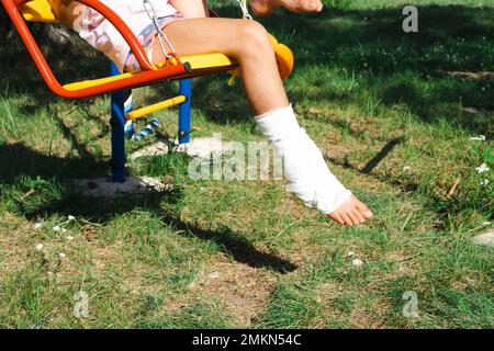 Kinderbeine sind mit Pflaster verbunden - barfuß auf einer Schaukel. Traumatische Ausrüstung, Kinderverletzung, Bluterguss, Verstauchung, Fraktur Stockfoto