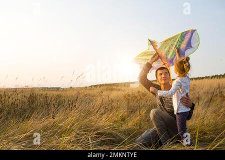 Papa hilft seiner Tochter, im Sommer bei Sonnenuntergang einen Drachen auf einem Feld zu fliegen. Familienunterhaltung im Freien, Vatertag, Kindertag. Ländliche Gebiete, zusatzbereich Stockfoto