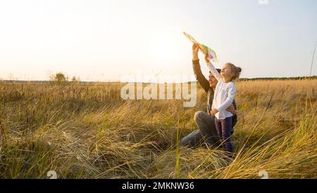 Papa hilft seiner Tochter, im Sommer bei Sonnenuntergang einen Drachen auf einem Feld zu fliegen. Familienunterhaltung im Freien, Vatertag, Kindertag. Ländliche Gebiete, zusatzbereich Stockfoto