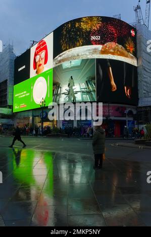 Ein Mann, der auf dem nassen Bürgersteig mit der berühmten Leuchtwerbung in Piccadilly steht. Zirkus in der Abenddämmerung in LONDON England Stockfoto