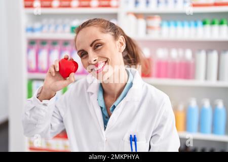 Junge, wunderschöne hispanische Frau, die Apotheke lächelt und ein Herz hält Stockfoto