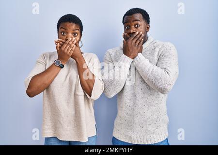 Ein junges afroamerikanisches Paar, das über blauem Hintergrund stand, schockierte den Mund mit Händen für einen Fehler. Geheimes Konzept. Stockfoto