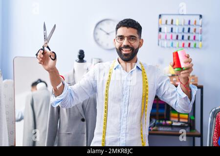 Ein junger arabischer Schneider, der selbstbewusst Schere und Faden in der Schneiderei hält Stockfoto