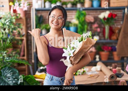 Eine junge asiatische Frau im Blumenladen, die einen Blumenstrauß hält und den Daumen zur Seite zeigt und glücklich mit offenem Mund lächelt Stockfoto