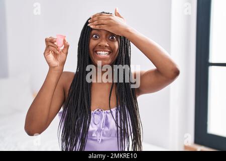 Junger afroamerikaner mit Zöpfen, die die Menstruationstasse halten, gestresst und frustriert von Hand auf Kopf, überrascht und wütend im Gesicht Stockfoto