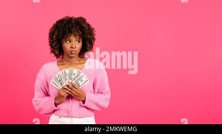 Behutsam fröhliche junge afro-amerikanische, lockige Frau in lässiger Kleidung hat eine Menge Geld und schaut auf leeren Raum Stockfoto