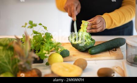 Die junge afroamerikanische Dame in der Schürze schneidet Brokkoli am Tisch mit Gemüse in der weißen Küche, aus nächster Nähe Stockfoto