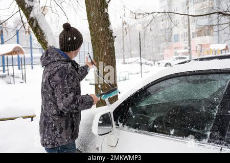 Ein Mann putzt Schnee aus einem Auto nach einem Schneefall. Straßenverkehrssicherheit, schwierige Witterungsbedingungen im Winter Stockfoto