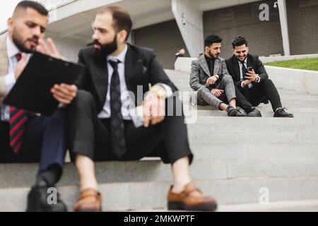 Geschäftstreffen im Freien. Auf den Stufen der Treppe sitzt eine Gruppe männlicher Geschäftsleute in Anzügen. Arbeitspause. Diskussion und Stockfoto