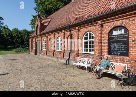 Ältere Frau, die auf einer weißen Holzbank vor einer roten Ziegelfassade im Garten von Schloss Rosenholm in Jütland, Dänemark sitzt Stockfoto