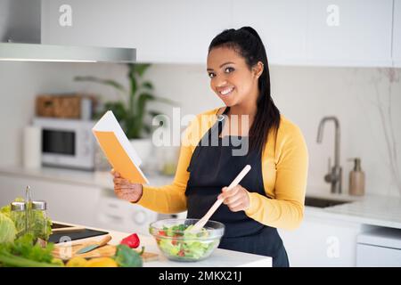 Neues Rezept und hausgemachtes Essen. Die junge afroamerikanische Dame in der Schürze macht Salat, liest Notizbuch Stockfoto