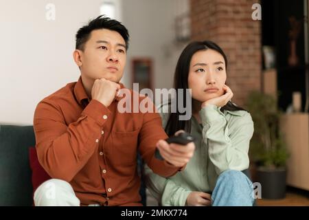 Unzufriedenes asiatisches Paar, das Fernsehen guckte, die Fernbedienung zeigte und die Fernsehkanäle wechselte, auf dem Sofa saß Stockfoto