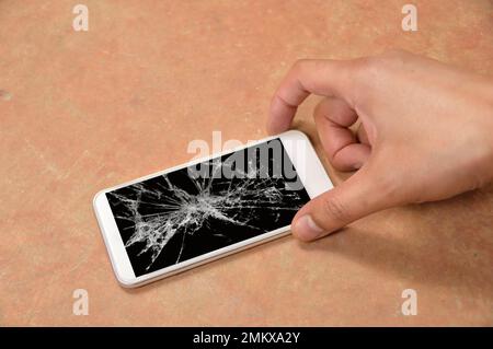 Person, die zu Hause ein kaputtes Smartphone mit einem rissigen Bildschirm auf dem Boden aussucht Stockfoto