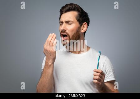 Mundgeruch. Ein gutaussehender Mann mittleren Alters, der seinen Atem mit der Hand kontrolliert, ihm bläst, über grauem Hintergrund steht Stockfoto