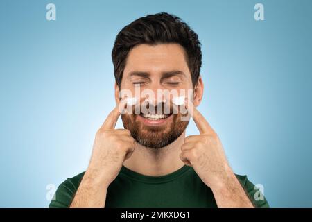 Fröhlicher, bärtiger, gutaussehender Mann, der Gesichtscreme auf die Wangen aufträgt, mit geschlossenen Augen und vor blauem Studiohintergrund posiert Stockfoto