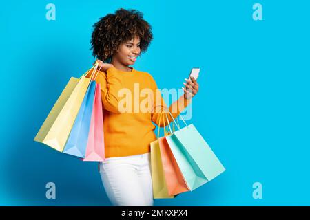 Positive afrikanische Frau, die online einkauft, Smartphone auf Blau benutzt Stockfoto