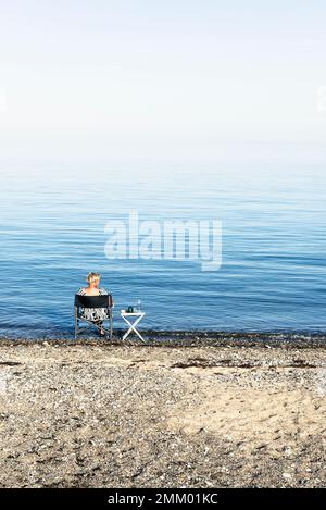 Eine blonde Frau sitzt auf einem Campingstuhl am Ufer von Kattegatt in der Sonne und trinkt Wein, Djursland, Jütland, Dänemark Stockfoto