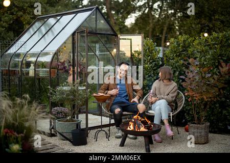 Ein junges Paar, das Spaß während eines romantischen und stimmungsvollen Abendessens am Kamin im Hinterhof mit altem Glashaus dahinter hat Stockfoto