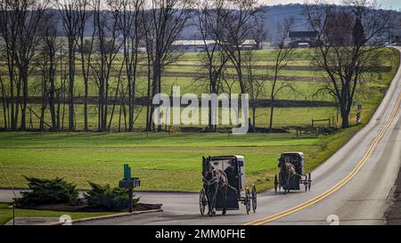 Ein Blick auf zwei Amish-Pferde und Buggys, die an einem sonnigen Dezember-Tag durch eine Landstraße durch die Felder fahren, und ein Schulhaus mit einem Zimmer Stockfoto