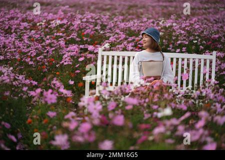 Lächelnde junge Chinesin sitzt auf einer Bank, umgeben von rosa Galsang-Blumen im Garten Stockfoto