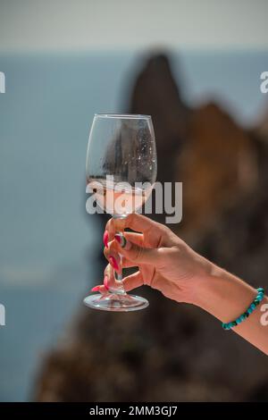 Eine weibliche Hand hält ein Glas Wein vor dem Hintergrund des Meeres und der Berge. Stockfoto