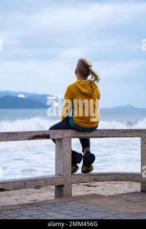 Ein einsamer Teenager, der am Wasser auf dem Meer sitzt. Rückansicht eines Teenagers gegen Meereslandschaft. Stockfoto