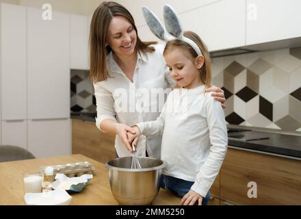 Mutter und ihre Vorschultochter in Hasenohren, die in der modernen Küche Teig zubereiten und schwingen. Lächeln und Spaß haben, zusammen zu sein. Osterkonzept Stockfoto