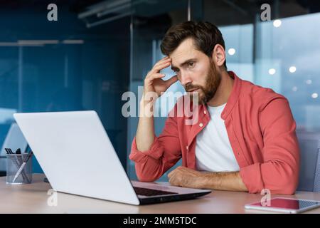 Ein gestresster und müder junger Geschäftsmann, Student, Freiberufler in einem roten Hemd sitzt im Büro an einem Laptop. Er hält seinen Kopf. Überstunden, Deadline, Probleme. Stockfoto