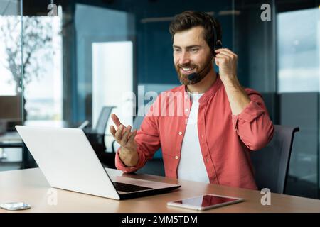 Porträt eines jungen Mannes mit Headset und rotem Hemd. Er sitzt am Schreibtisch im Büro, arbeitet an einem Laptop, kommuniziert per Videoanruf, spricht, lächelt. Stockfoto