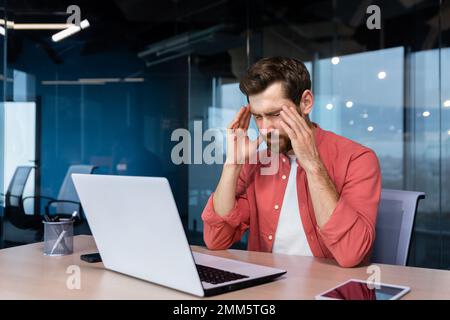 Stress und Kopfschmerzen am Arbeitsplatz. Ein junger Geschäftsmann in einem roten Hemd sitzt am Schreibtisch im Büro, hält seinen Kopf und schließt die Augen. Er fühlt sich schlecht, macht sich Sorgen um Probleme, ein Projekt, Überstunden, Stockfoto