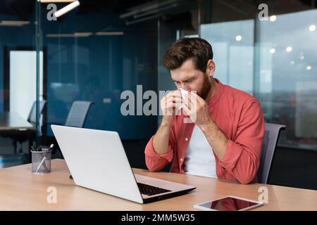 Ein junger Mann ist krank auf der Arbeit. Sitzt am Schreibtisch im Büro, wischt sich die Nase mit einer Serviette, hat eine laufende Nase, fühlt sich nicht gut an. Stockfoto