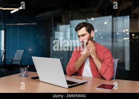 Ein junger Mann, ein Student, ein Freiberufler sitzt am Schreibtisch im Büro. Studiert, arbeitet auf einem Laptop. Er hält seine Wange, hat starke Zahnschmerzen. Stockfoto