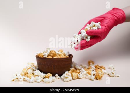 Abstrakte Zusammensetzung. Popcorn-Mix in einer Keramikschüssel. Popcorn läuft von der Hand in die Schüssel. Auf der Hand ist eine medizinische Handschuhfarbe Viva Magenta. Stockfoto