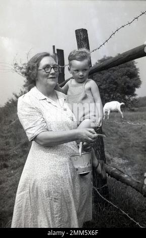 1950er, historisch, draußen auf dem Bauernhof, ein kleiner Junge in einem einteiligen Wollkostüm, der von seiner Oma gehalten wird, während sie an einem eingezäunten Feld steht, kurz davor, Ziegen auf einem eingezäunten Feld in England, Großbritannien, einen Leckerbissen zu schenken. Stockfoto