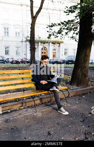 Stilvoller, lockiger Mann, der ein Buch liest, während er im Park sitzt Stockfoto
