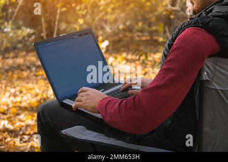 Ein freiberuflicher Mann arbeitet in der Natur im Herbstwald. Landleben. Eine Pause von der Zivilisation. Van Lifevibes. Ein Mann mit Laptop. Fernunterricht an der frischen Luft über einen Laptop. Remote-Arbeit. Stockfoto