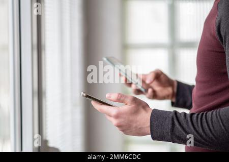 Nahaufnahme eines Mannes mit zwei Smartphones. Mann, der zwei Telefone gleichzeitig benutzt. Beschäftigter Mann, der neben Fenstern steht und zwei Smartphones benutzt. Stockfoto