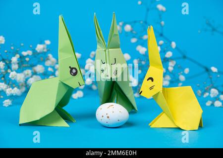 Drei Origami-Häschen aus Papier und ein Osterei auf blauem Hintergrund. Basteln Sie Ostern mit Ihren eigenen Händen Stockfoto