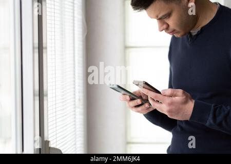 Nahaufnahme eines Mannes mit zwei Smartphones. Mann, der zwei Telefone gleichzeitig benutzt. Beschäftigter Mann, der neben Fenstern stand und zwei Smartphones benutzte. Stockfoto