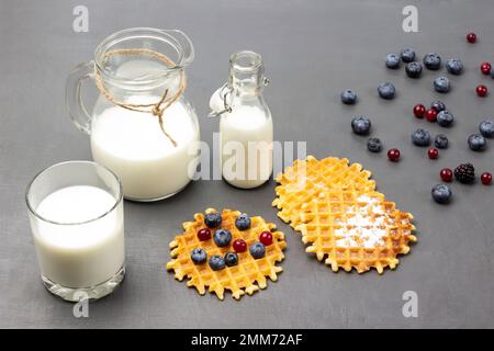 Waffeln und Beeren auf dem Tisch. Milch im Glas. Milch in Glasflasche und Kanne. Draufsicht. Grauer Hintergrund. Stockfoto