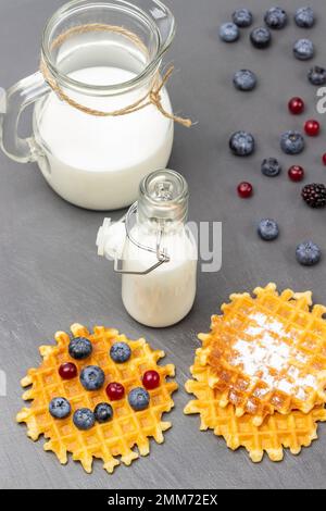 Waffeln und Beeren auf dem Tisch. Milch in Glasflasche und Kanne. Draufsicht. Grauer Hintergrund. Stockfoto