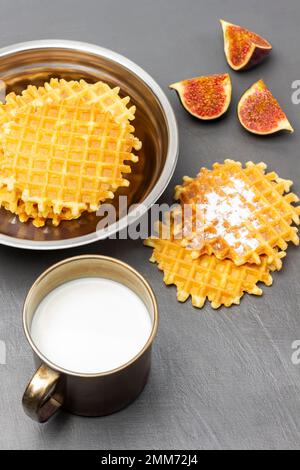 Wiener Waffeln in Metallschüssel. Waffeln und Feigenhälften auf dem Tisch. Milch in einem Metallbecher. Flach verlegt. Grauer Hintergrund. Stockfoto