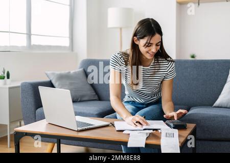 Junge Frau, die Haushaltssteuerrechnungen und -Ausgaben kalkuliert und zu Hause auf dem Sofa sitzt Stockfoto