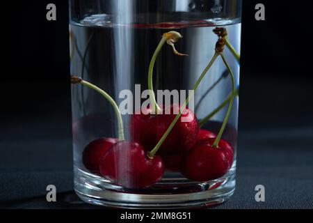 Kirschen in einem Glas Wasser auf schwarzem Hintergrund auf dem Tisch, Obst Hintergrund, Früchte Stockfoto