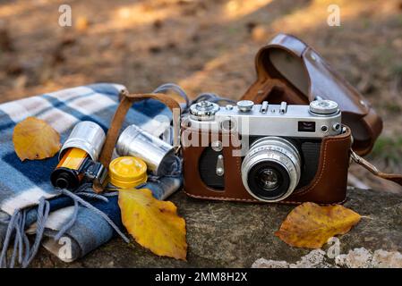 Oldtimer-Fotokamera in brauner Ledertasche und Fotofilm in einer Kartusche mit Schal und Herbstblättern auf einem Stein im Freien. Selektiver Fokus. Stockfoto