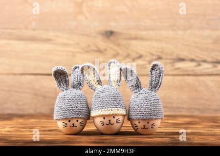 Drei Ostereier mit lustigen Gesichtern und graue Häkelhüte mit Hasenohren auf Holzhintergrund. Frohes Osterkonzept. Grußkarte. Stockfoto