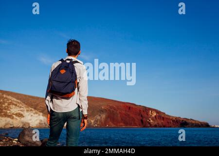 Reisender mit Rucksack, der die rote Strandlandschaft auf Santorini Island, Griechenland, mit Hügeln und Meer bewundert. Allein unterwegs. Sommerferien Stockfoto