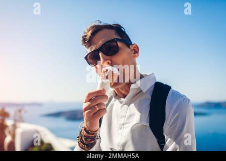 Ein Mann, der Eis in Zapfen auf der Insel Fira Santorini am Meereshintergrund von Caldera isst. Sommerferienessen. Schließen Stockfoto