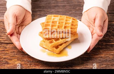 Köstliche belgische Waffeln mit Honig auf weißem Teller und Holzhintergrund, Frauenhände, rustikales Konzept. Stockfoto